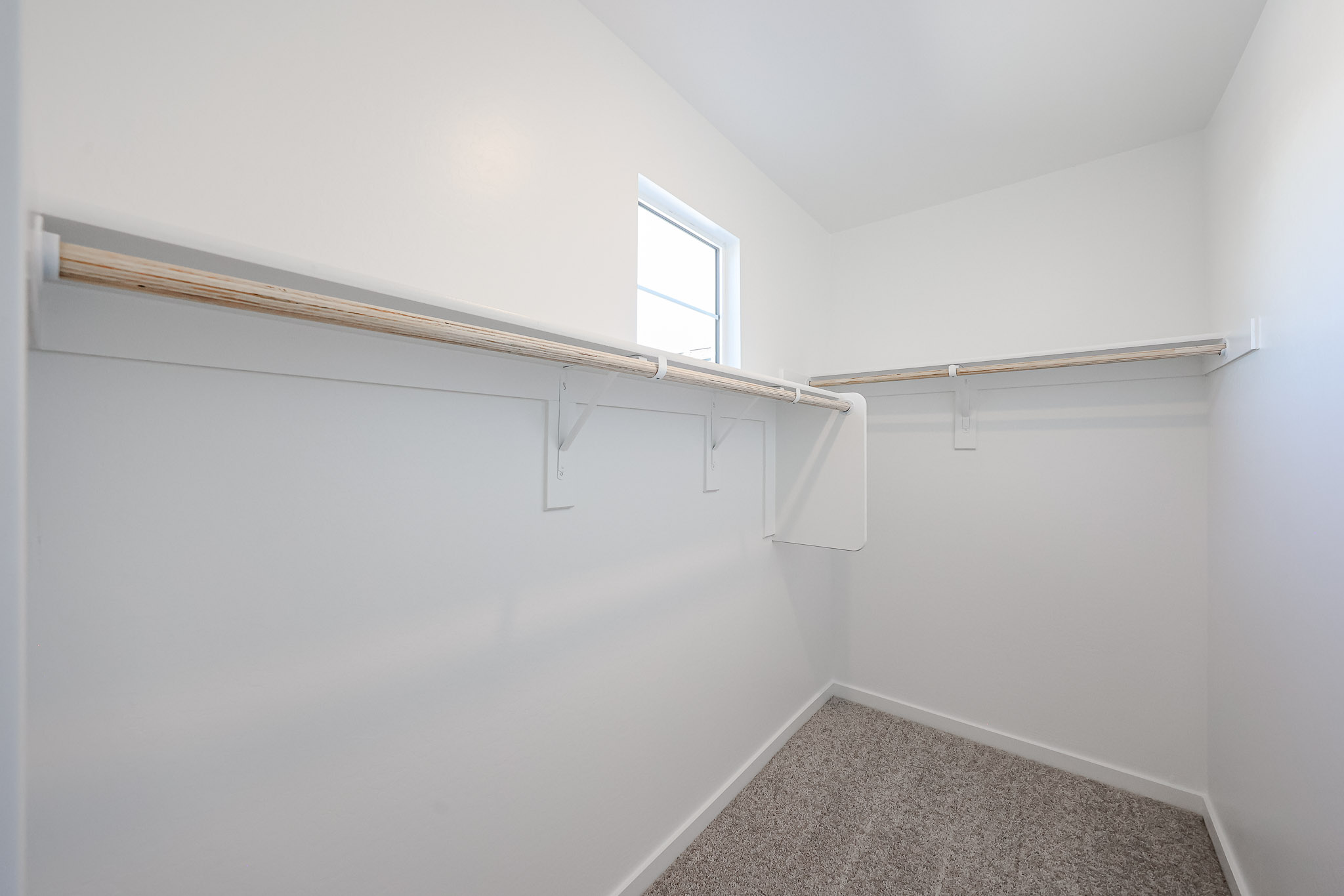 Empty walk-in closet with two wooden hanging rods, white walls, a small window, and carpeted floor.