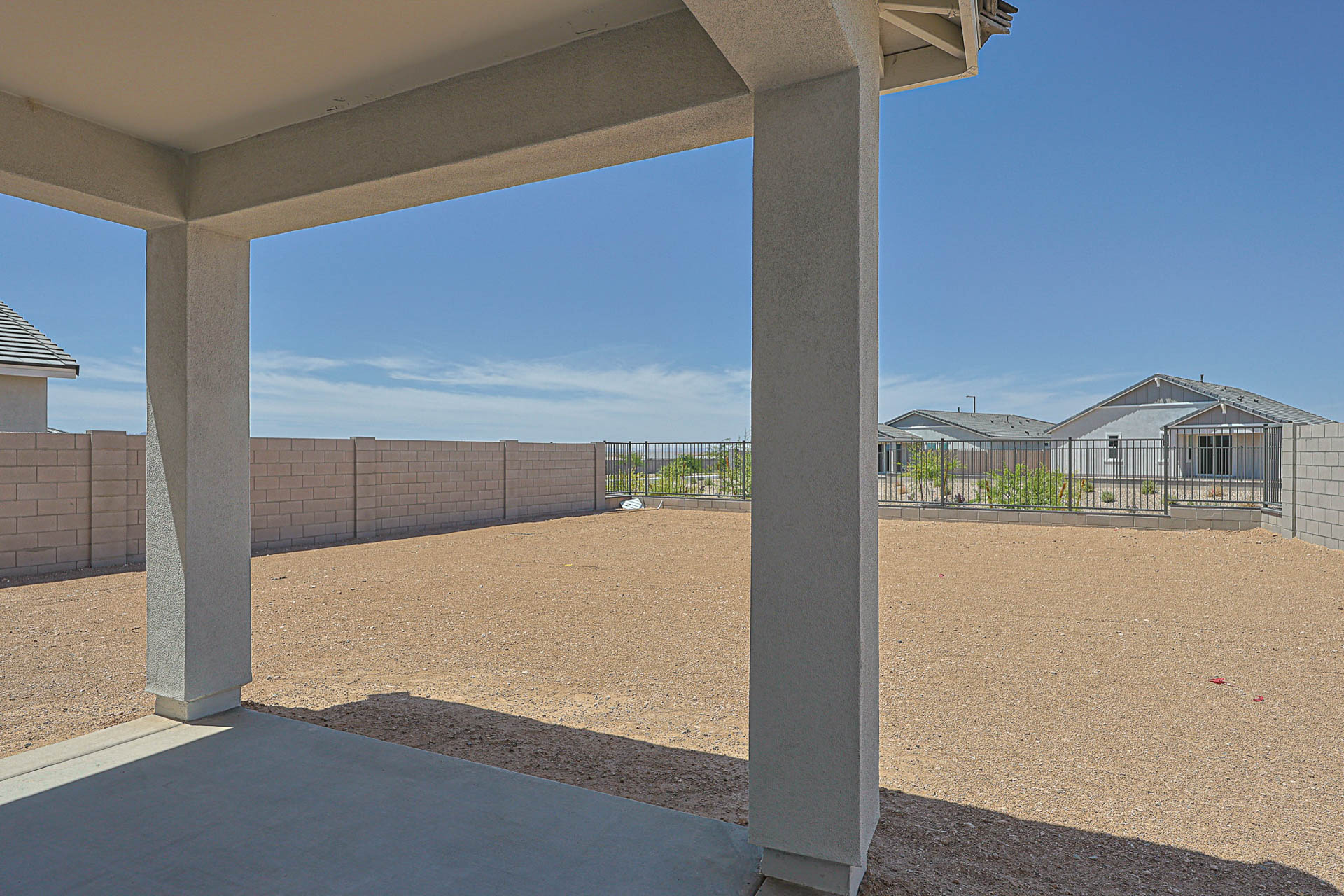 View of a covered patio looking out onto a large, fenced, dirt backyard with neighboring houses and a clear sky in the background.