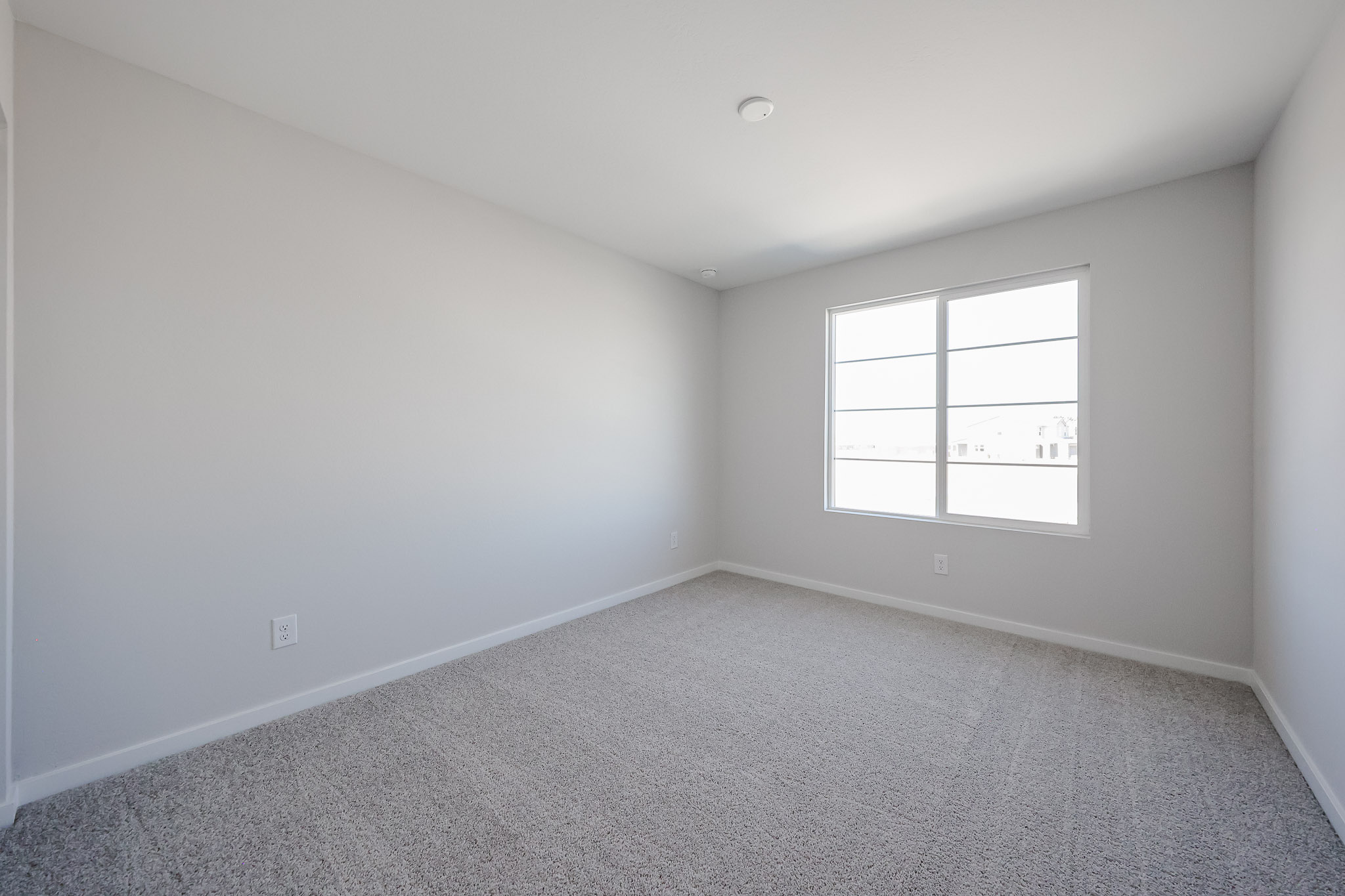 Empty room with light gray carpet, white walls, a large window letting in natural light, and a white ceiling. No furniture or decorations are present.