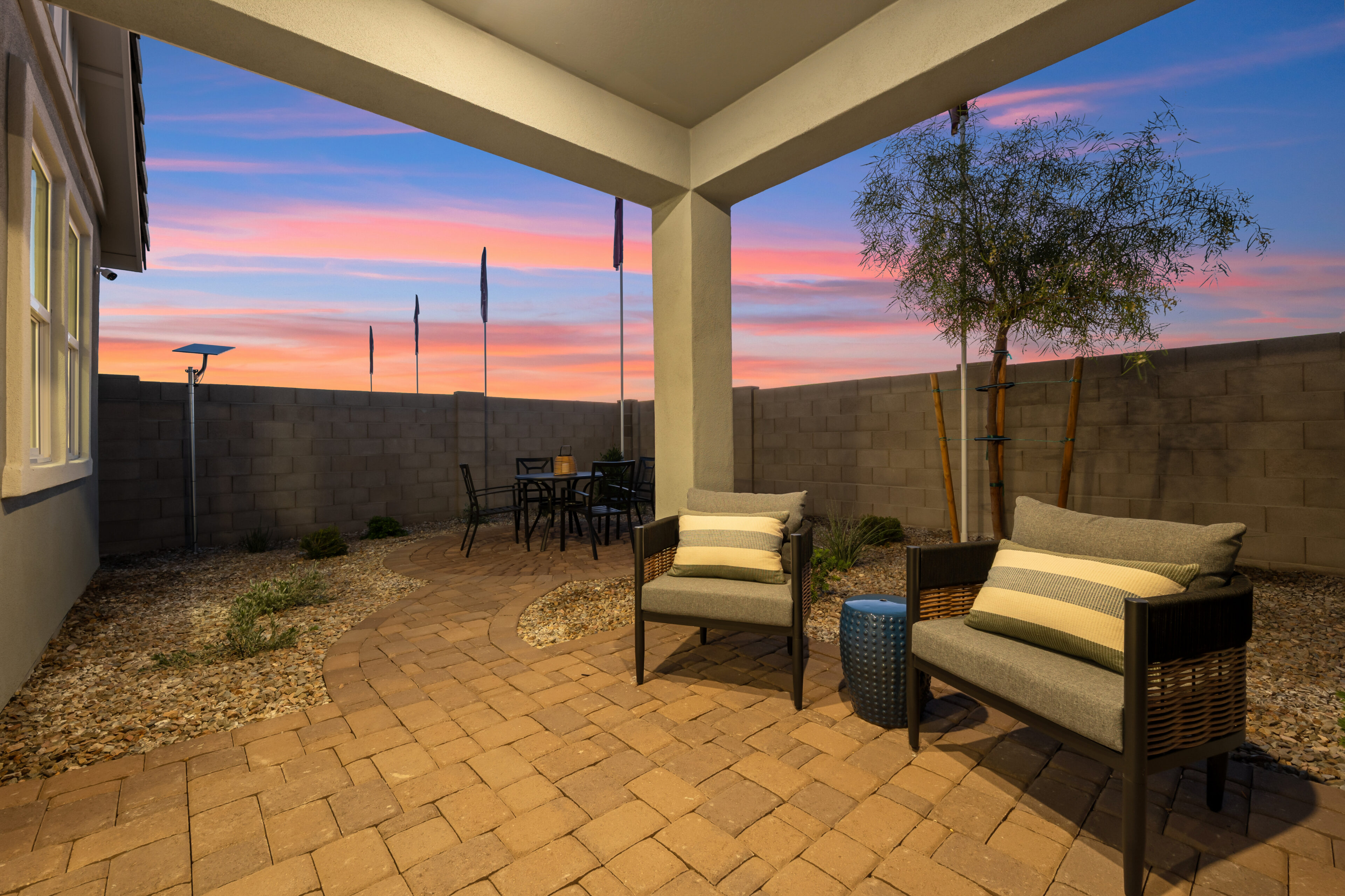 Covered patio with two chairs and a small table, brick flooring, outdoor dining set, gravel landscaping, and a sunset sky in the background.