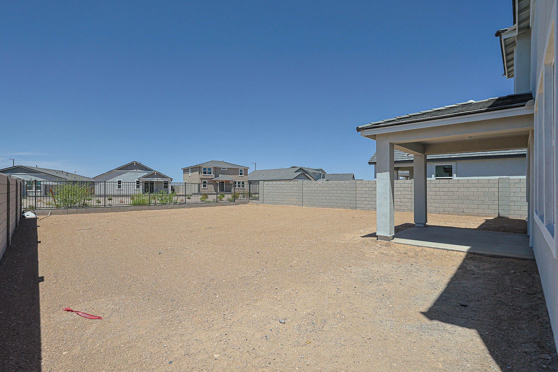 A backyard with bare dirt, a covered patio area, and a block wall fence. Neighboring houses are visible under a clear blue sky.