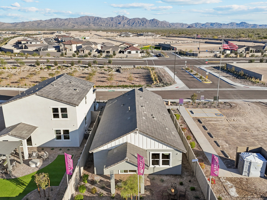 Aerial view of suburban houses with yards, wide streets, and desert landscape; American flag and mountain range visible in the background under a blue sky.