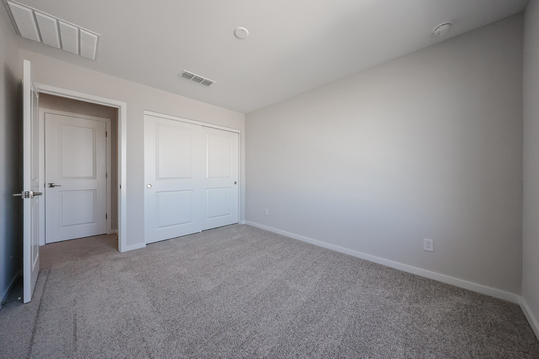 Empty room with beige carpet, light gray walls, a white closet with sliding doors, and a white entrance door. Ceiling vents and outlets are visible.