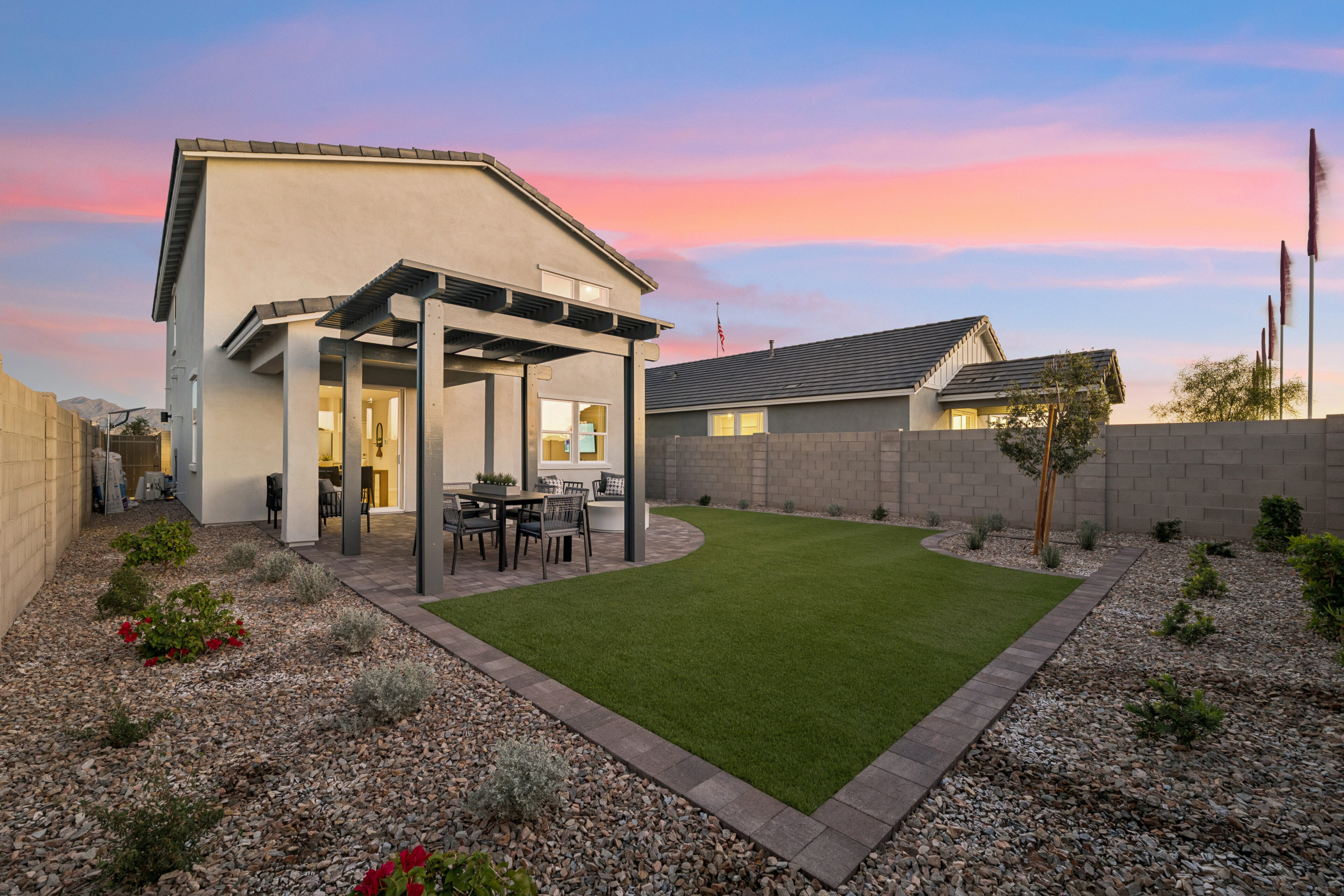 A modern backyard with artificial turf, a covered patio with outdoor seating, and landscaped gravel areas, surrounded by concrete block walls at sunset.