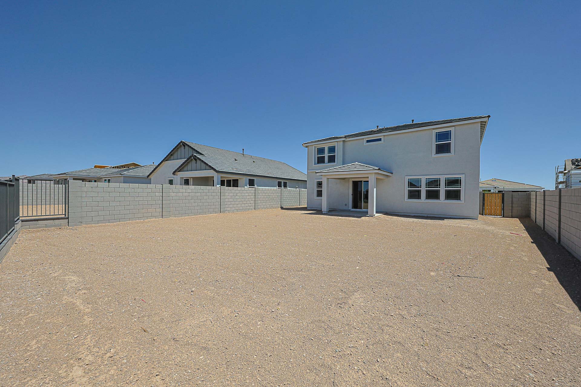 A two-story white house with multiple windows stands in a large, empty dirt backyard enclosed by a gray block wall under a clear blue sky.