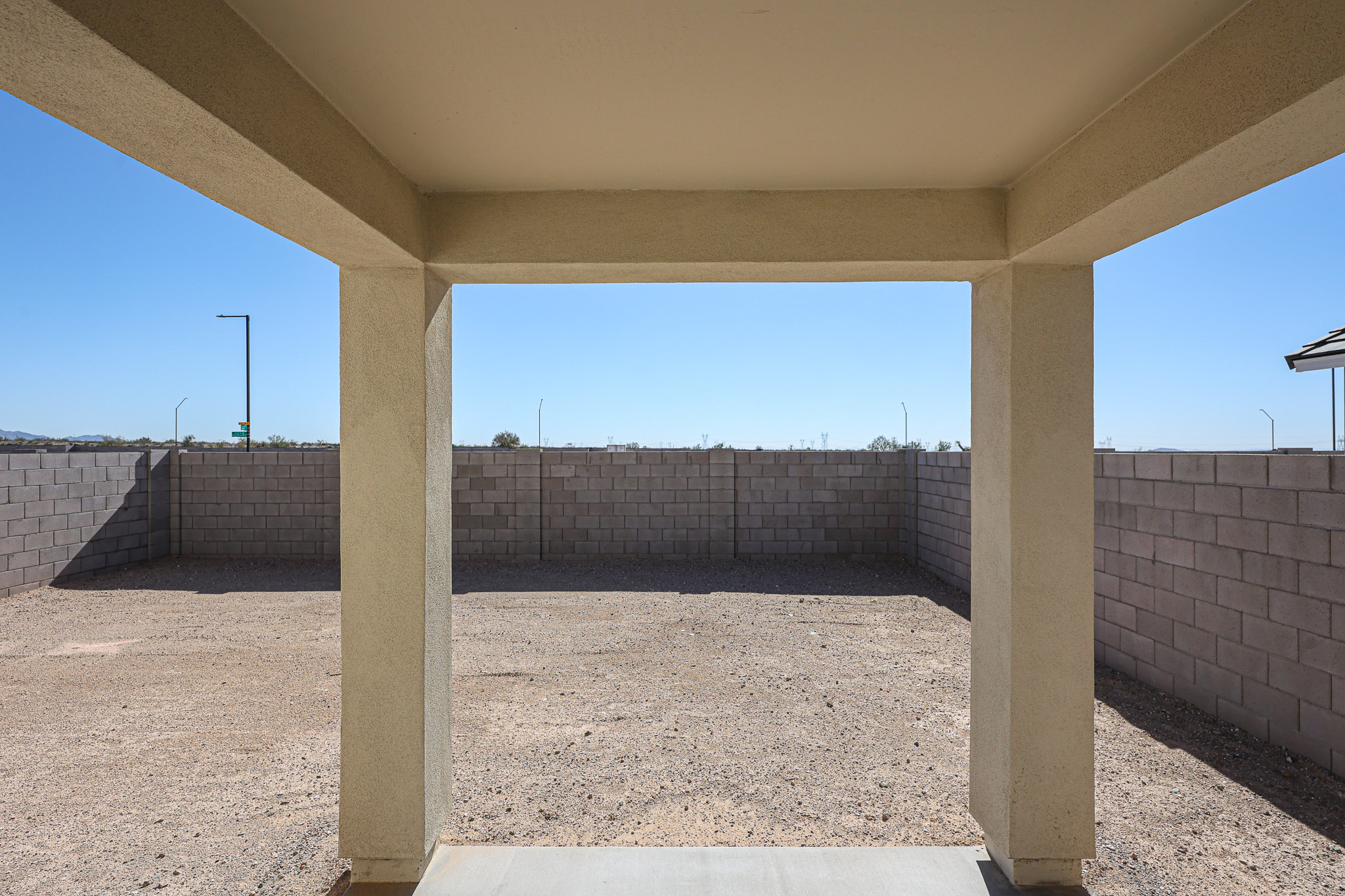View from a covered patio looking out onto a bare, fenced backyard with a gravel surface and concrete block walls under a clear blue sky.