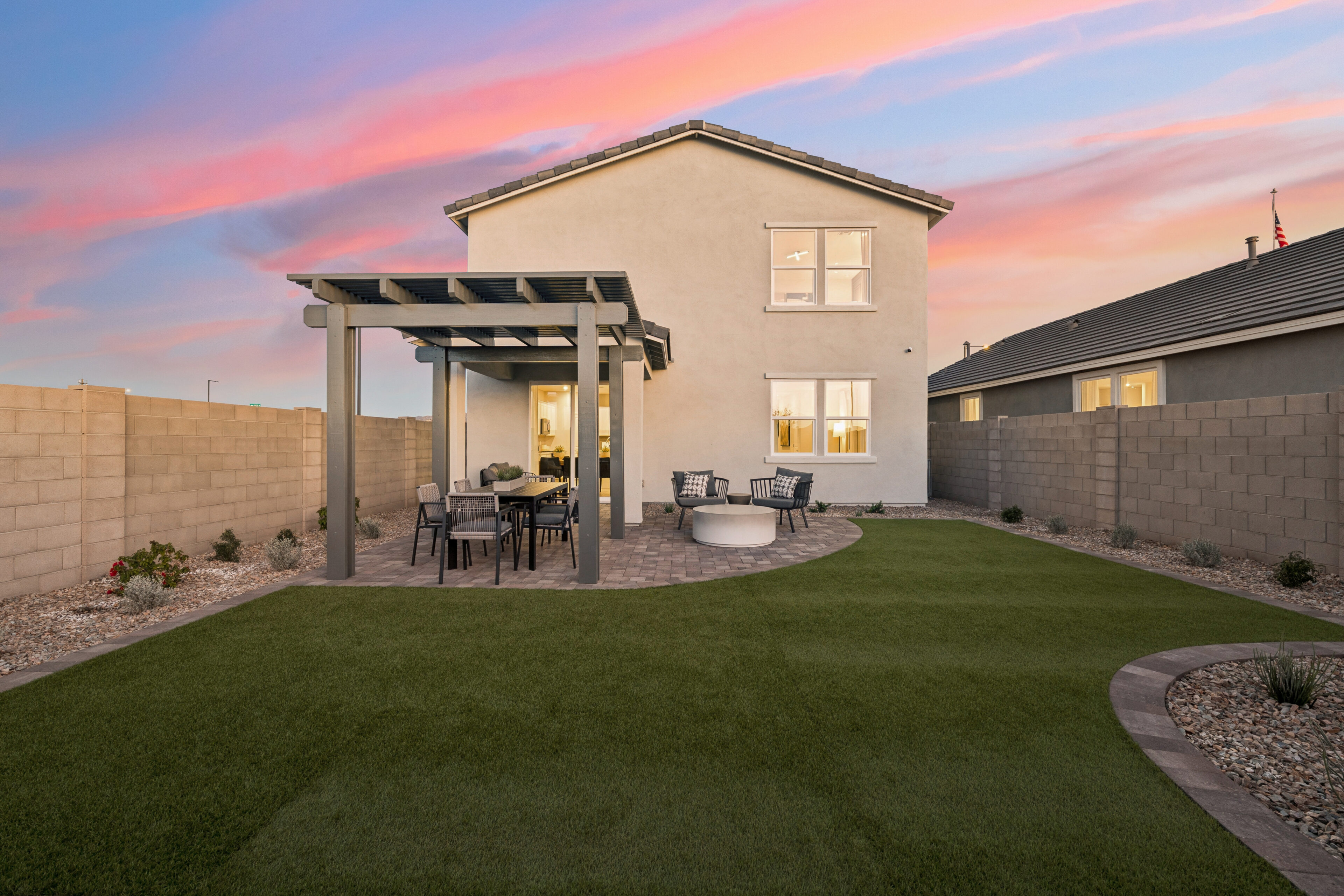 A modern backyard features artificial grass, a patio with a pergola, outdoor furniture, a fire pit, and is enclosed by a block wall, with a two-story house at sunset.