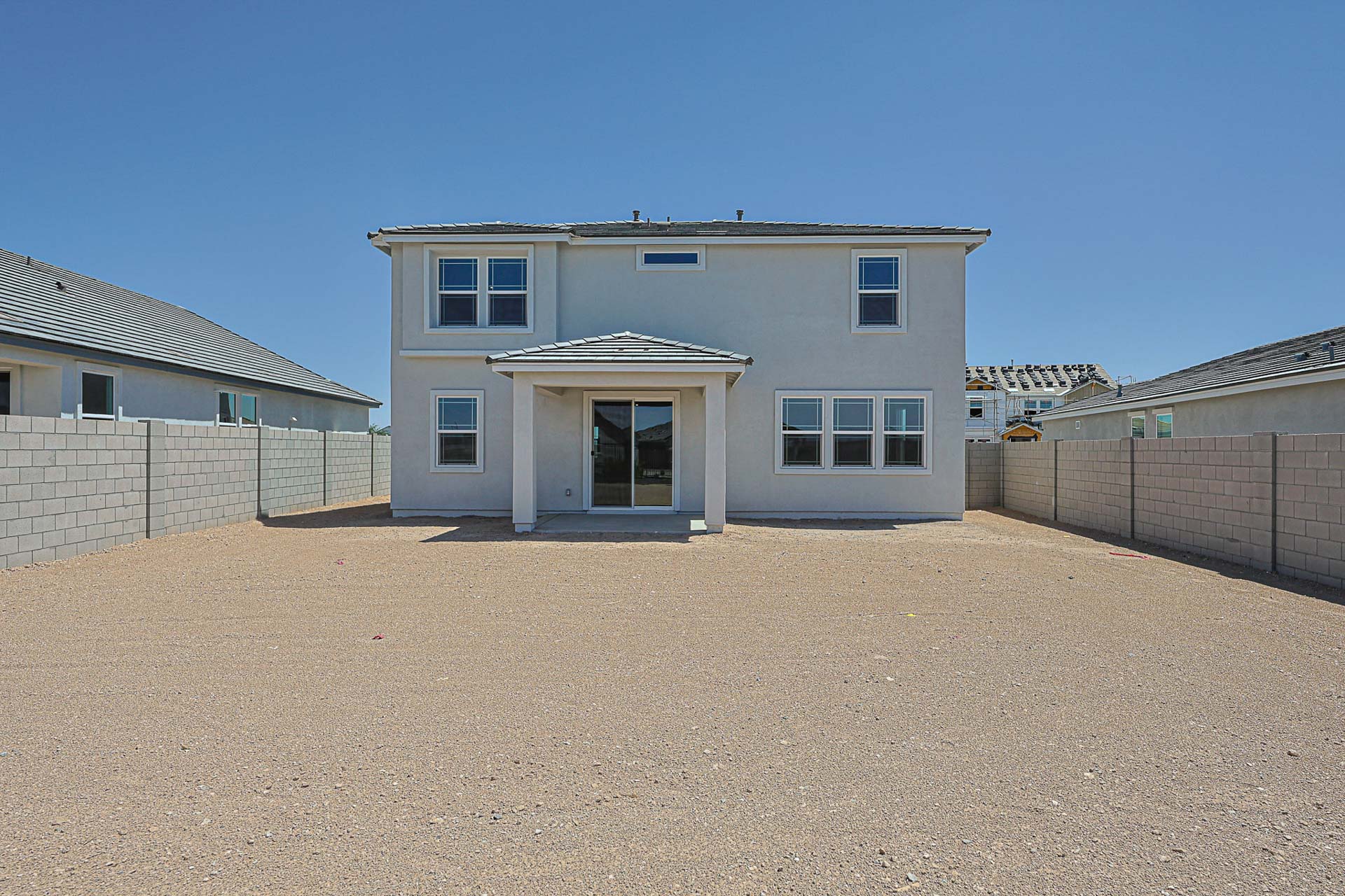 Two-story white house with a covered patio, surrounded by a gravel backyard and a gray block wall on both sides, under a clear blue sky.