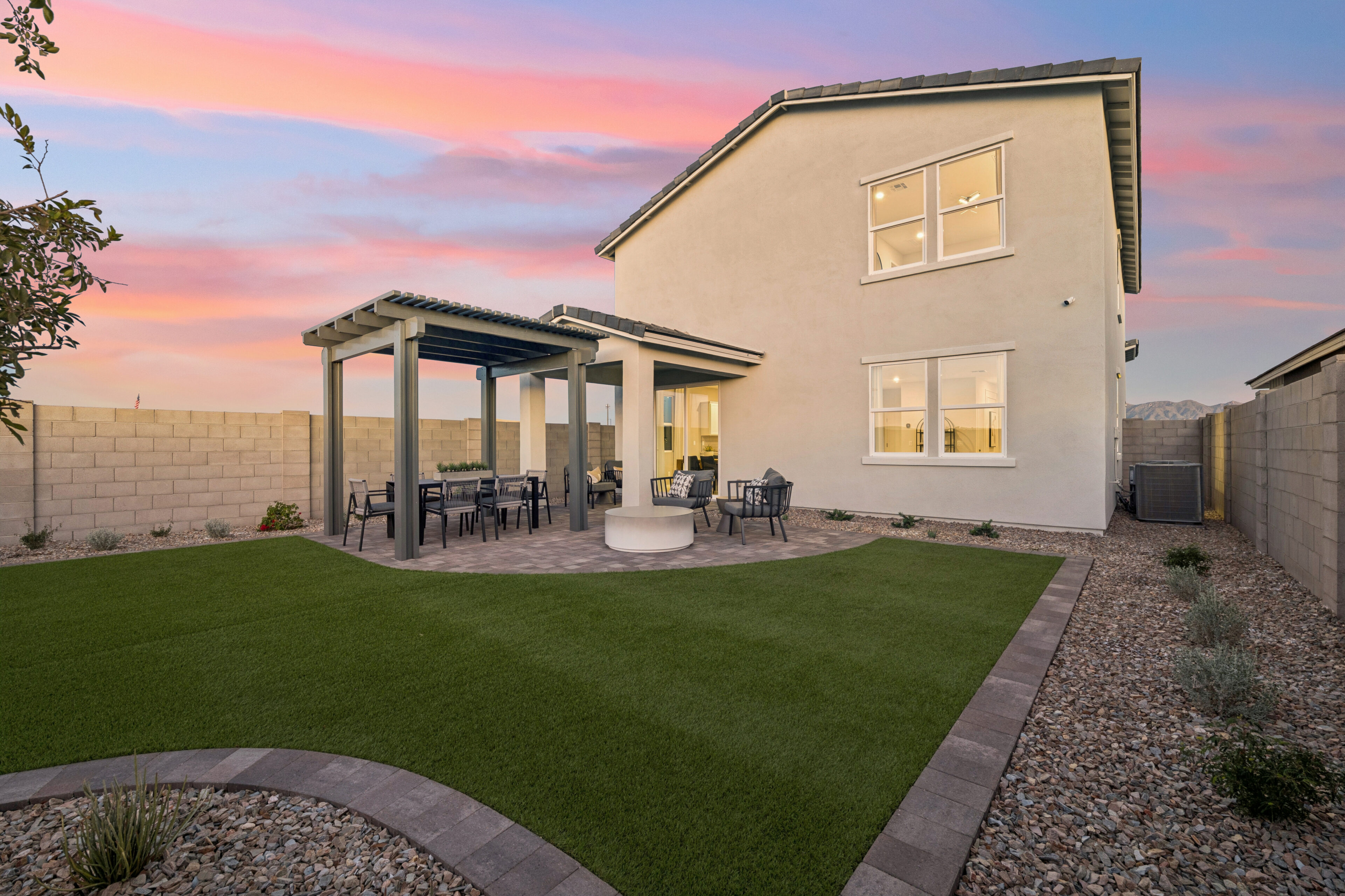 A modern backyard with artificial grass, a covered patio area, outdoor seating, a fire pit, and a stone pathway, enclosed by a high block wall under a colorful sunset sky.