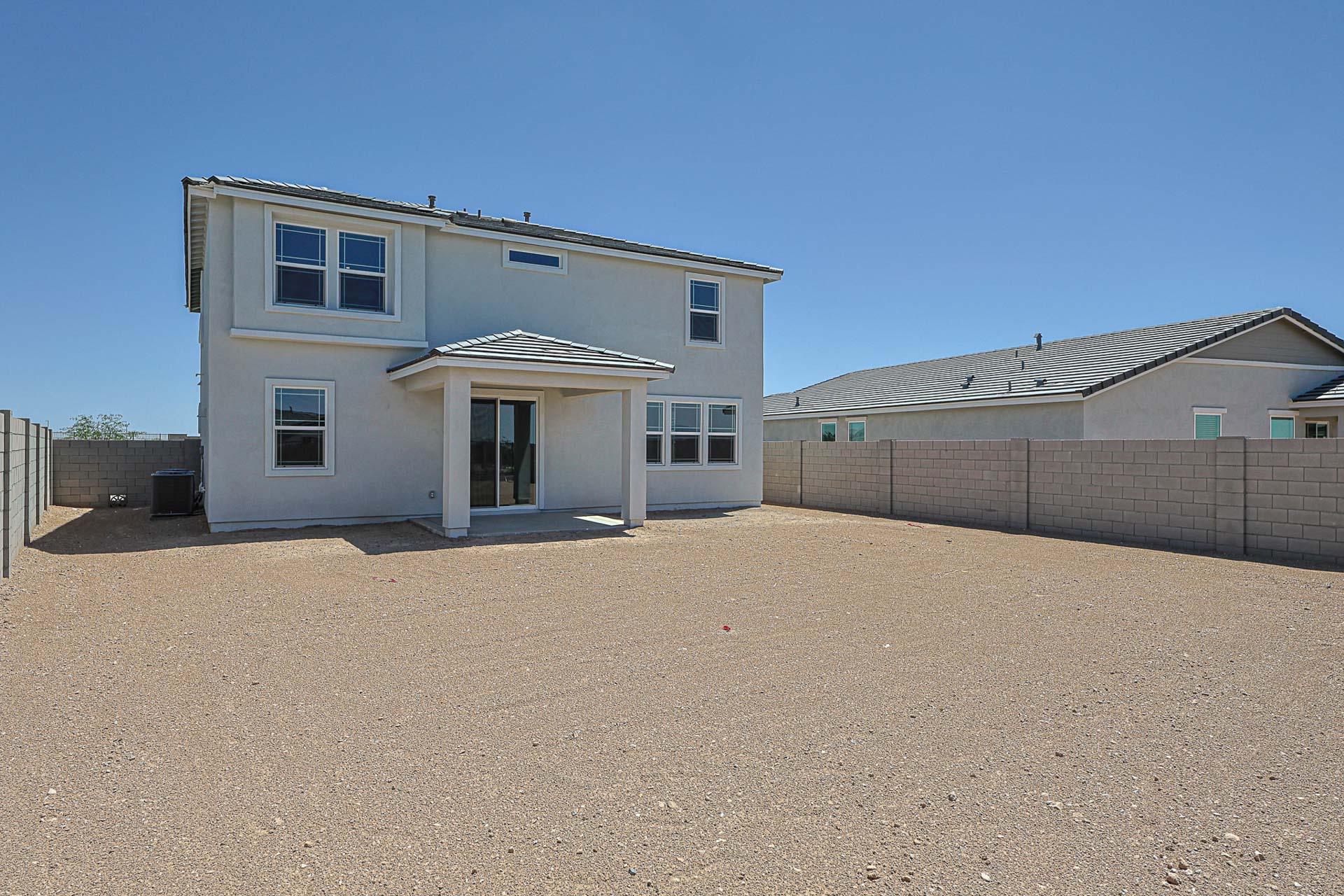 Two-story house with a covered patio and gravel backyard enclosed by a concrete block wall. Sky is clear and blue.
