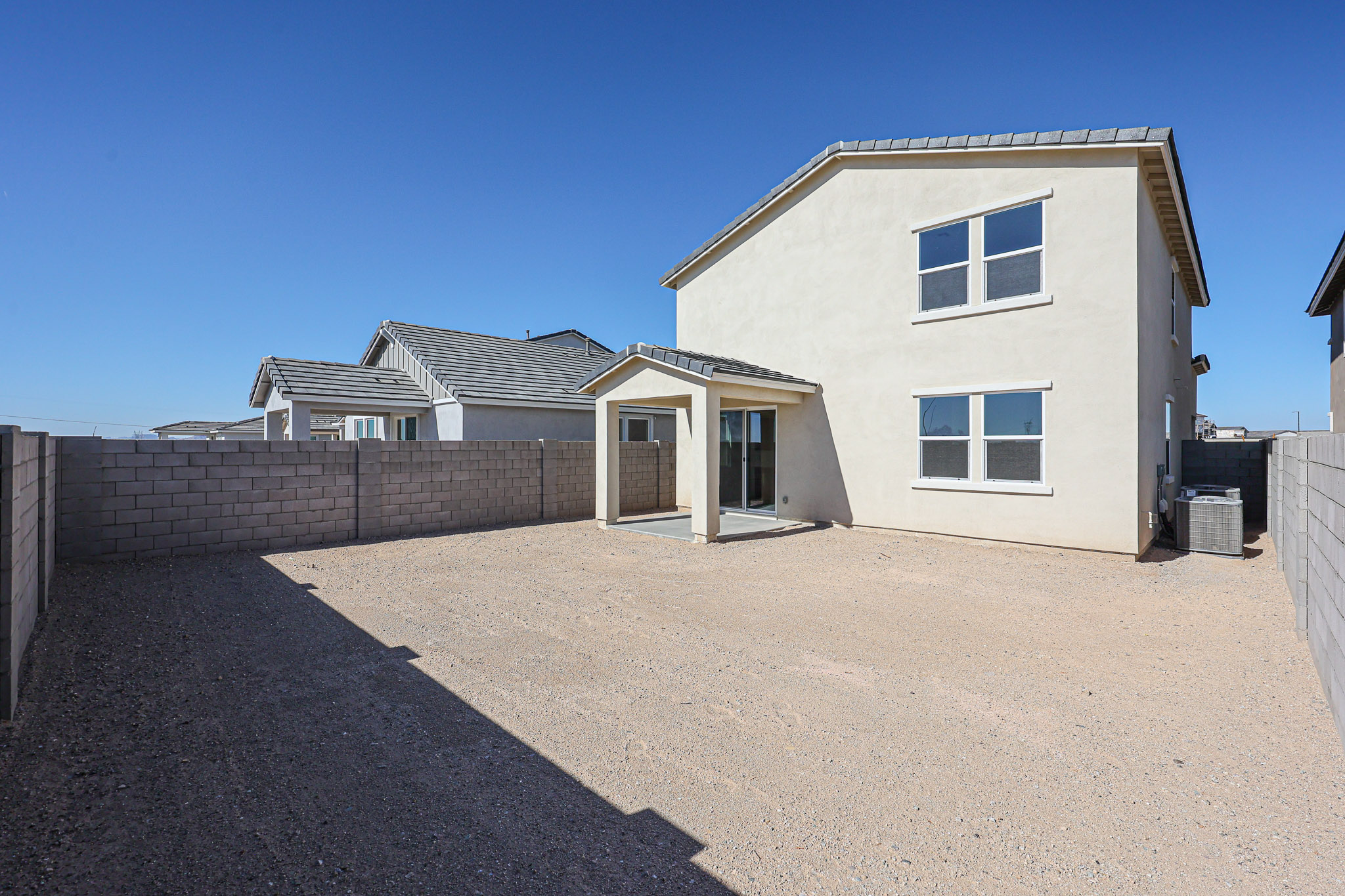 A modern two-story house with a small covered patio and a large, gravel backyard enclosed by a concrete block wall under a clear blue sky.