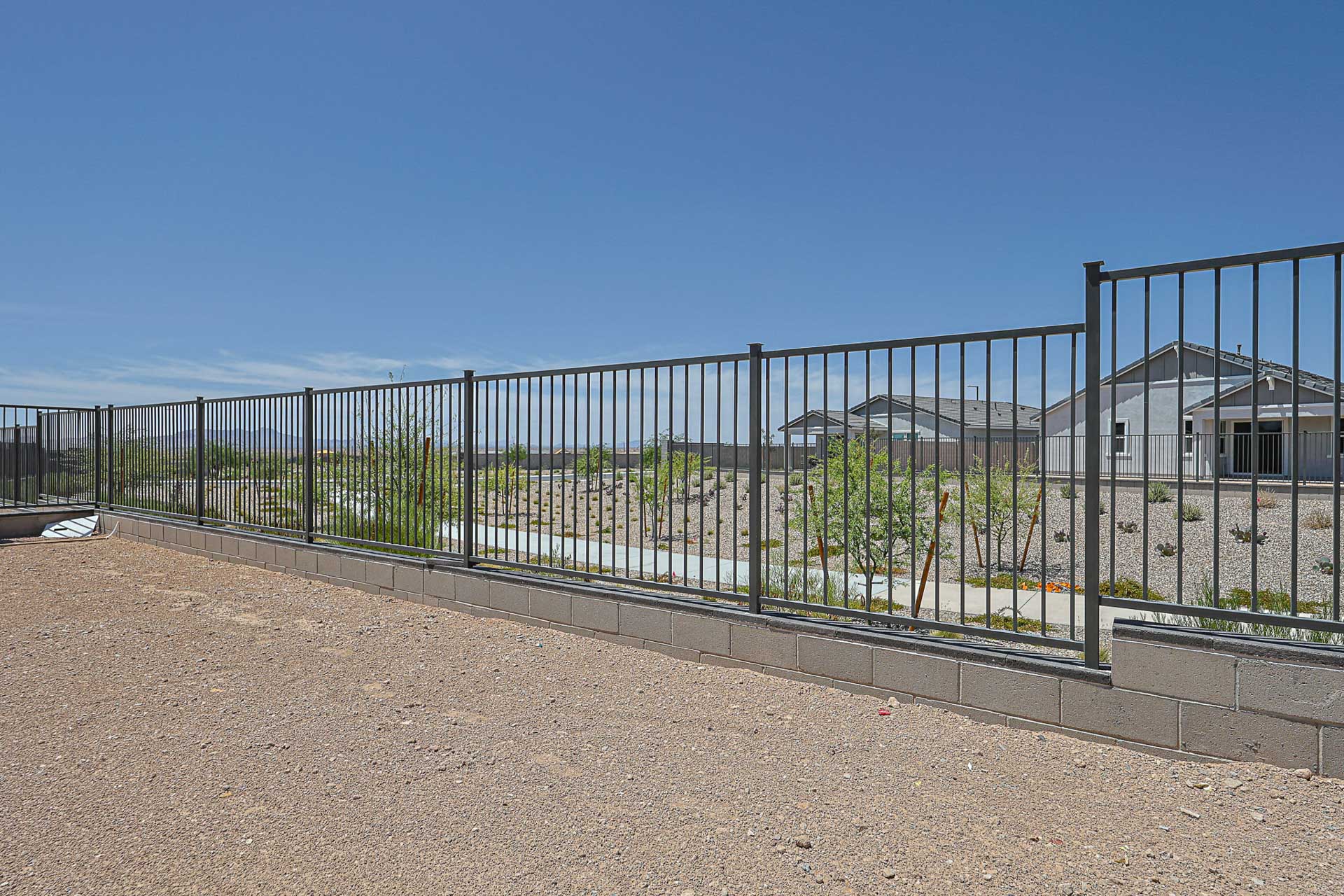 A metal fence with vertical bars stands on a low concrete block wall, separating a gravel yard from a residential street and houses under a clear blue sky.