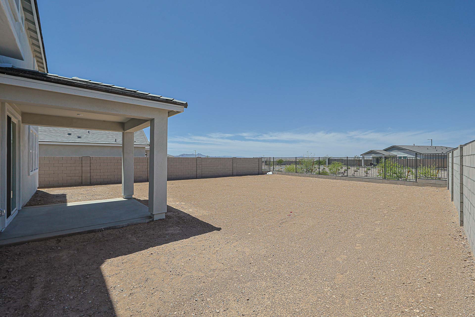 A fenced backyard with gravel ground, a covered patio area attached to a house, and a clear view of distant mountains under a blue sky.
