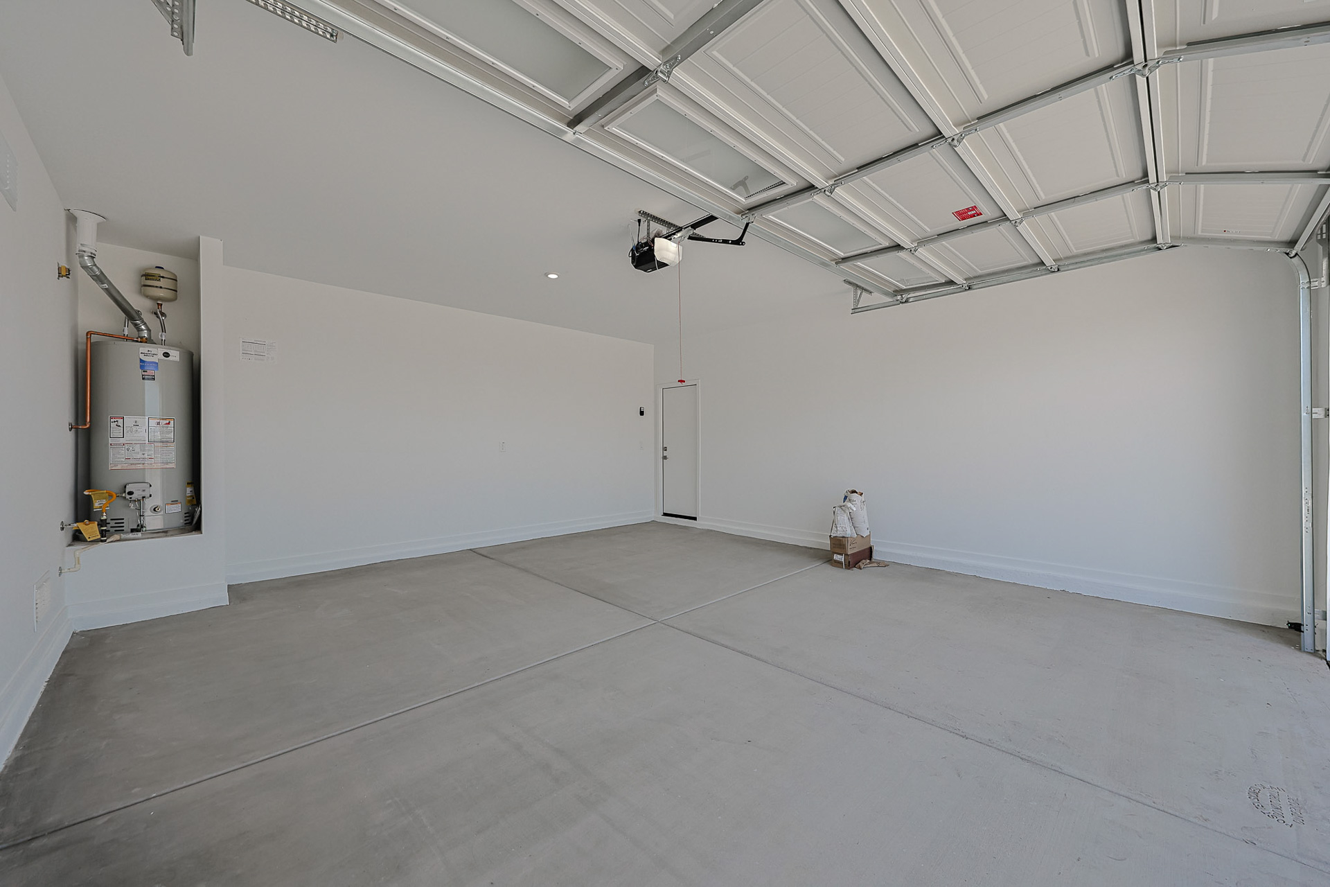 Empty two-car garage with a concrete floor, white walls, ceiling-mounted opener, water heater in the corner, and a small box near the back door.