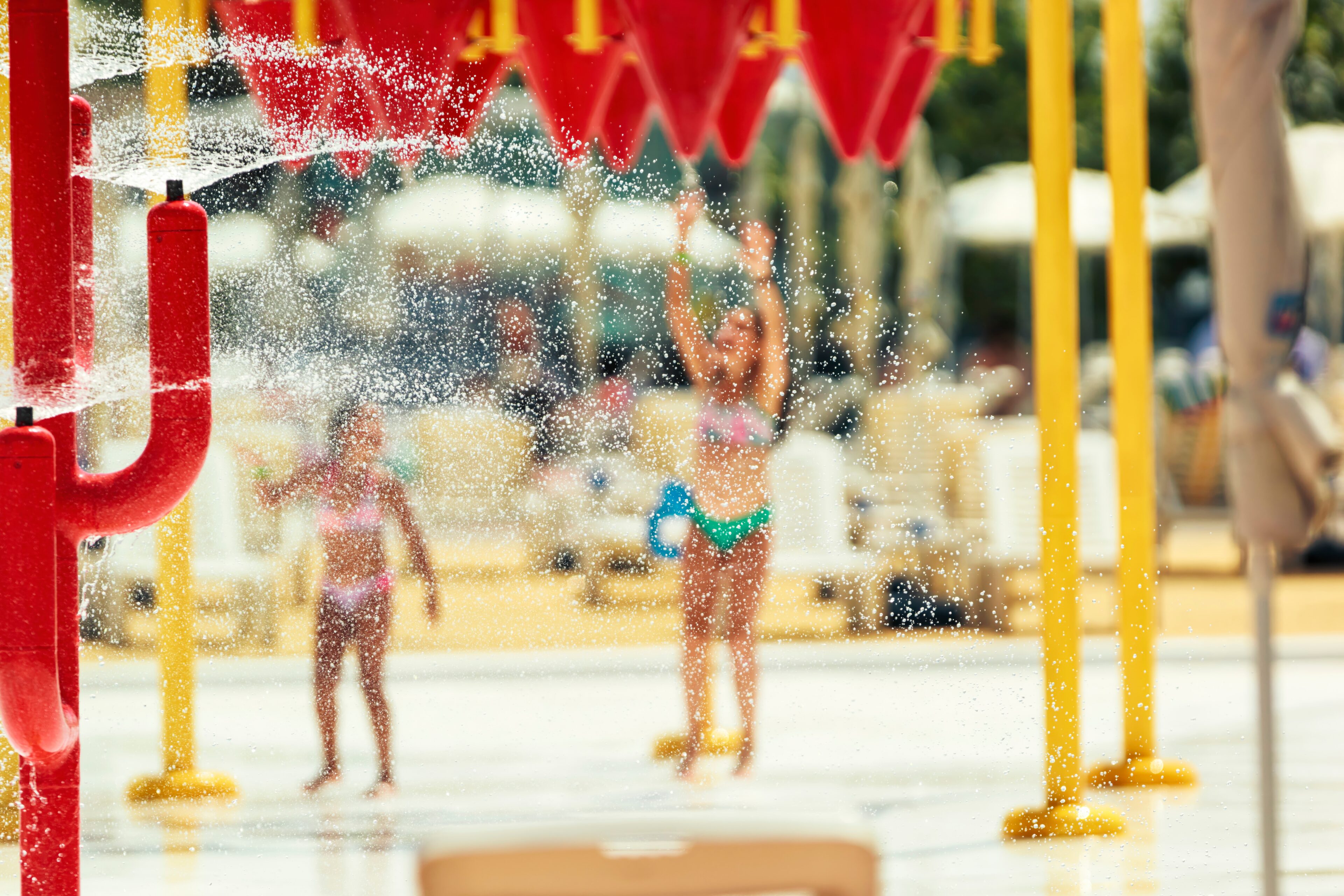 Children play in a splash pad area as water sprays from red and yellow structures on a sunny day.