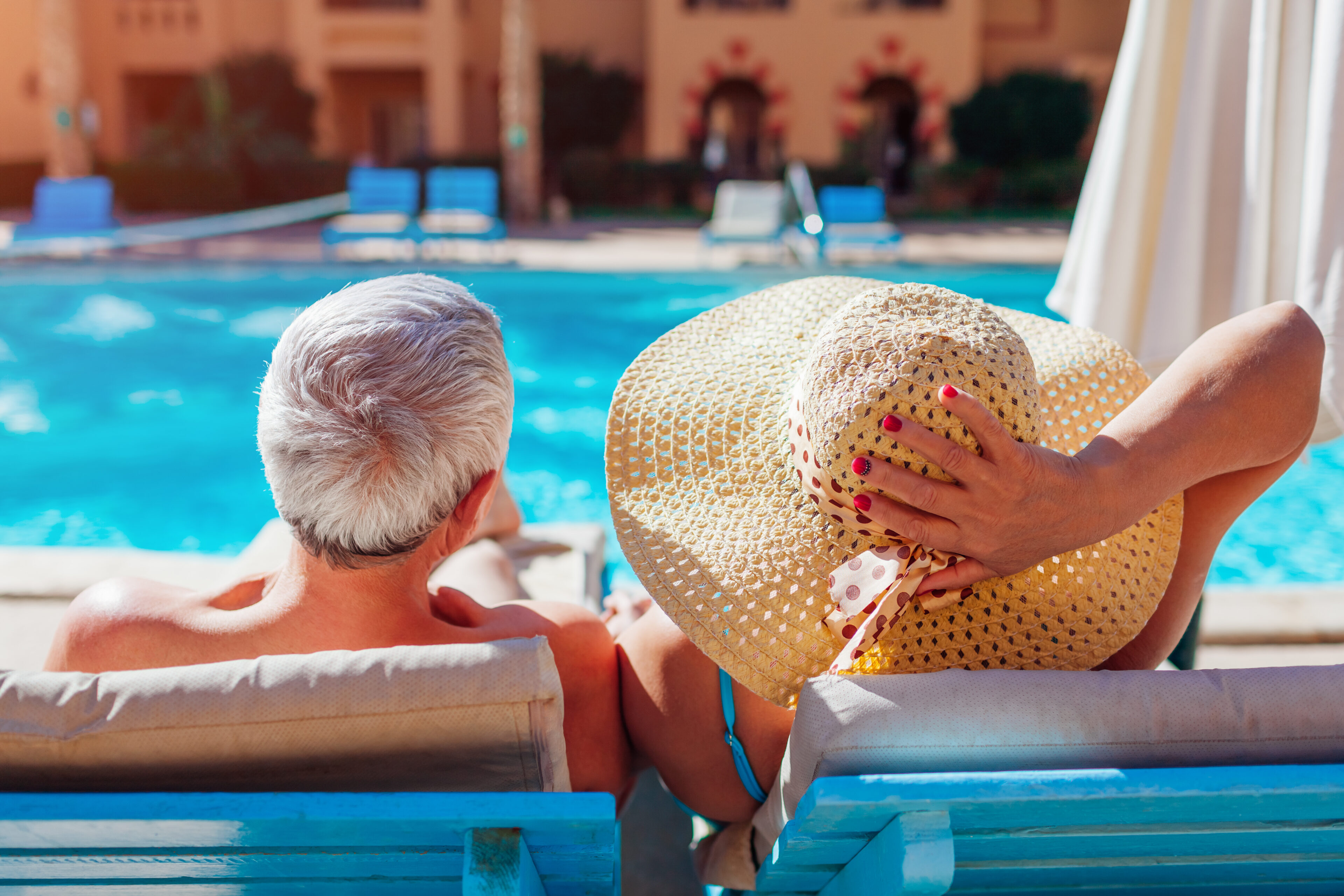 Two people sit side by side on lounge chairs by a swimming pool, with one wearing a wide-brimmed hat, facing away from the camera.