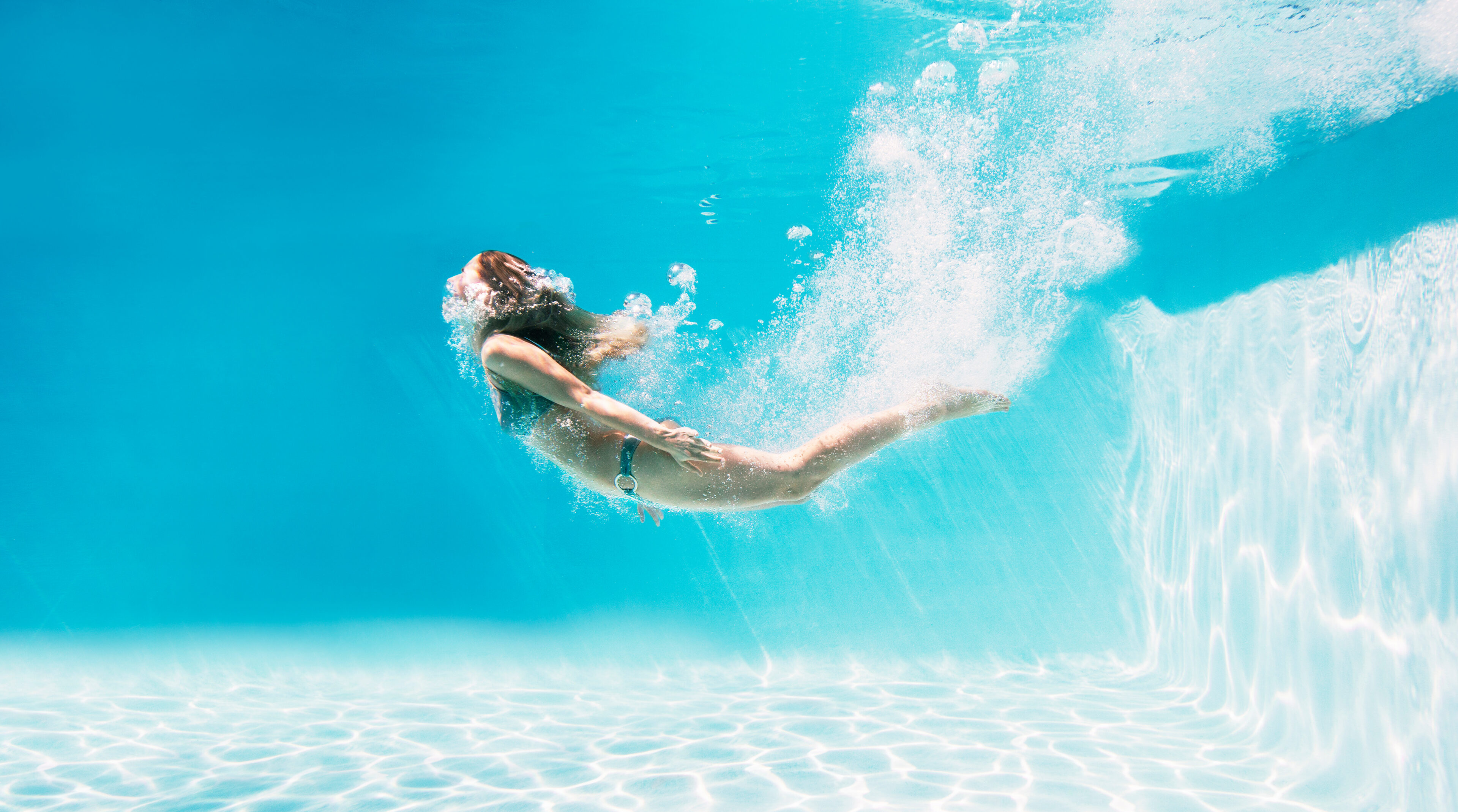 Person in a swimsuit swims underwater in a pool, blowing bubbles and creating a splash near the pool floor and wall.