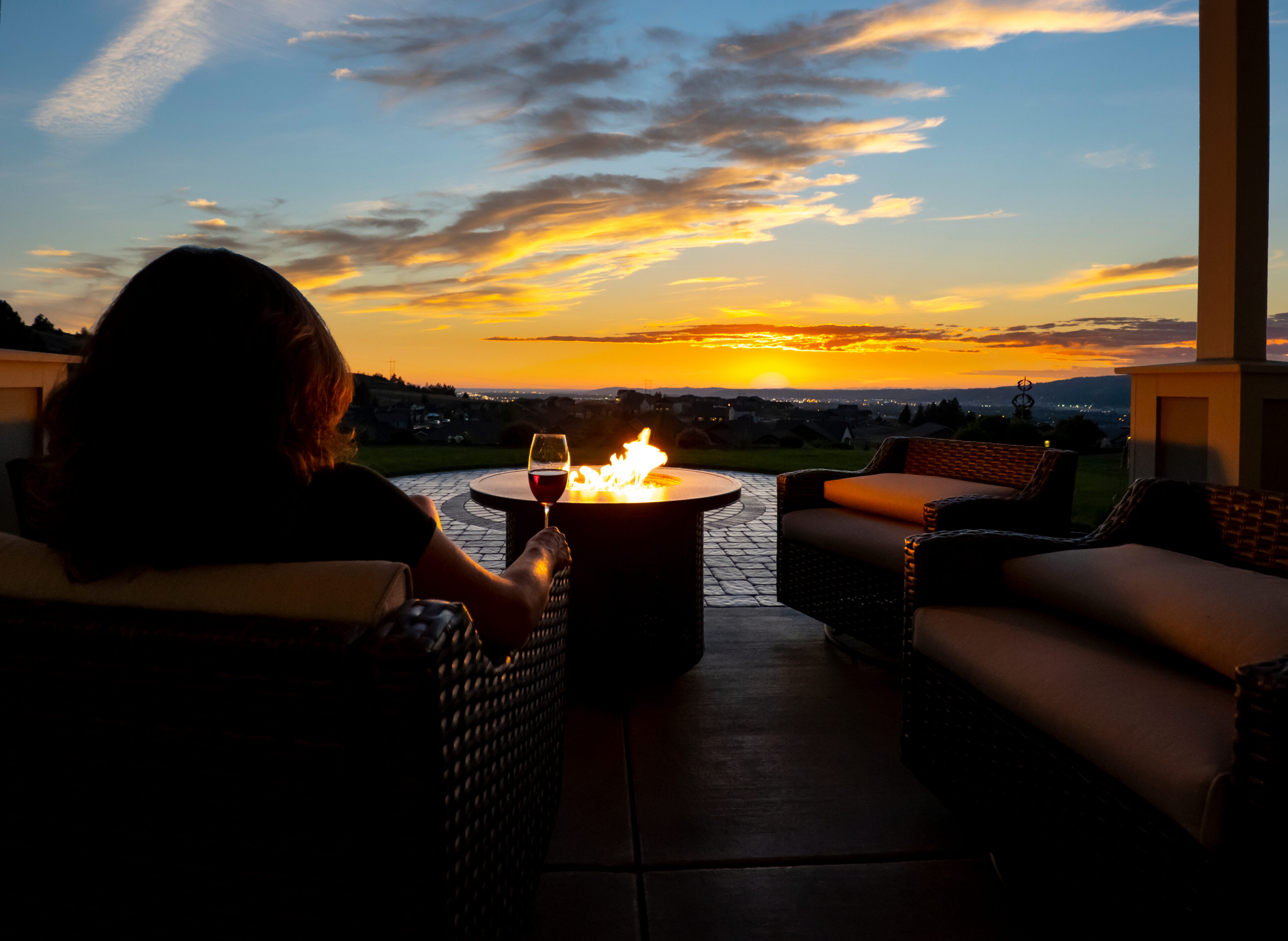 Person sits on a patio chair by a fire pit with a glass of wine, overlooking a scenic sunset and open landscape.