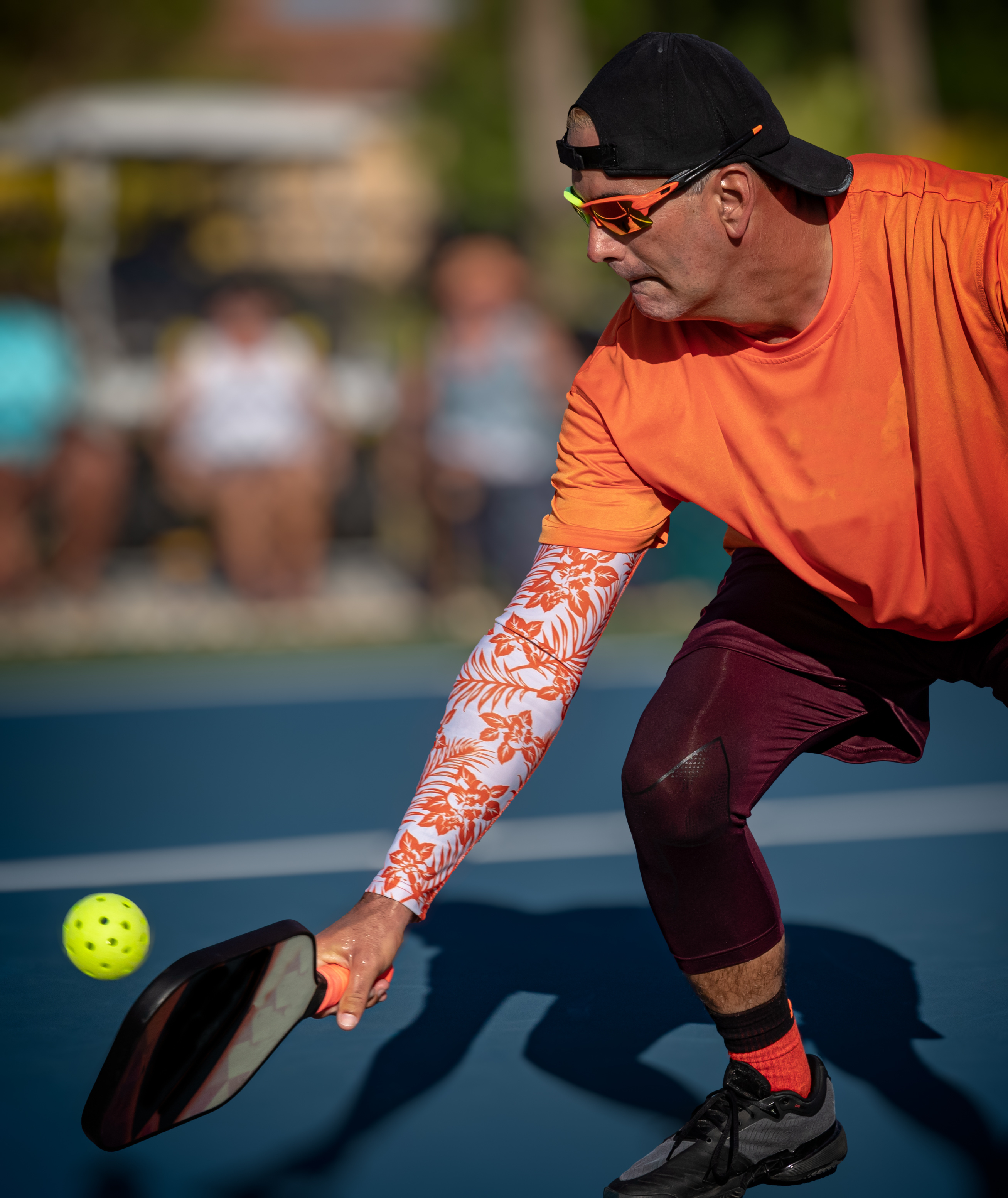 A man in an orange shirt and cap plays pickleball, reaching low with his paddle to hit a yellow ball on an outdoor court.
