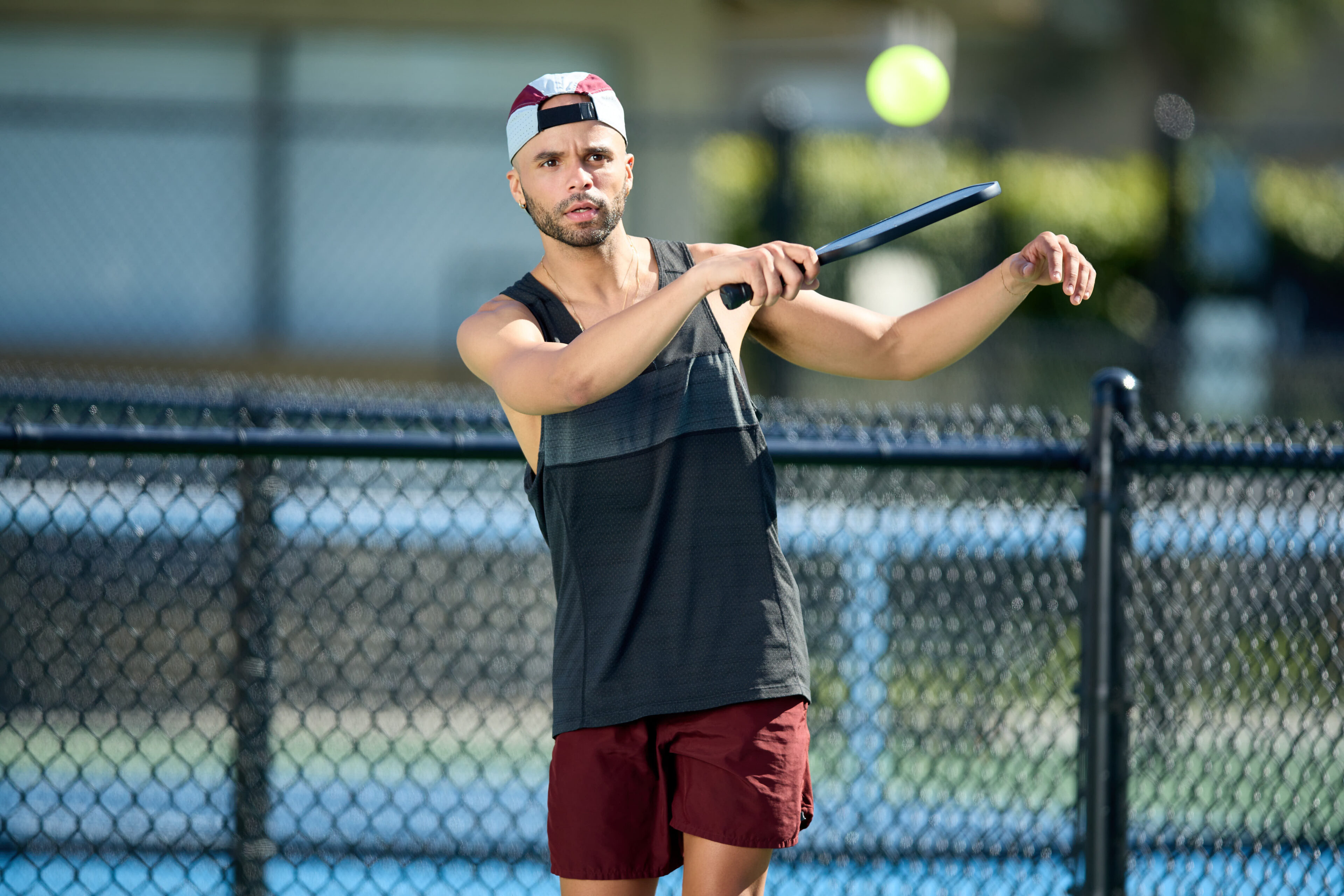 A man in athletic clothing and a backwards cap prepares to hit a tennis ball with a racquet on an outdoor court.