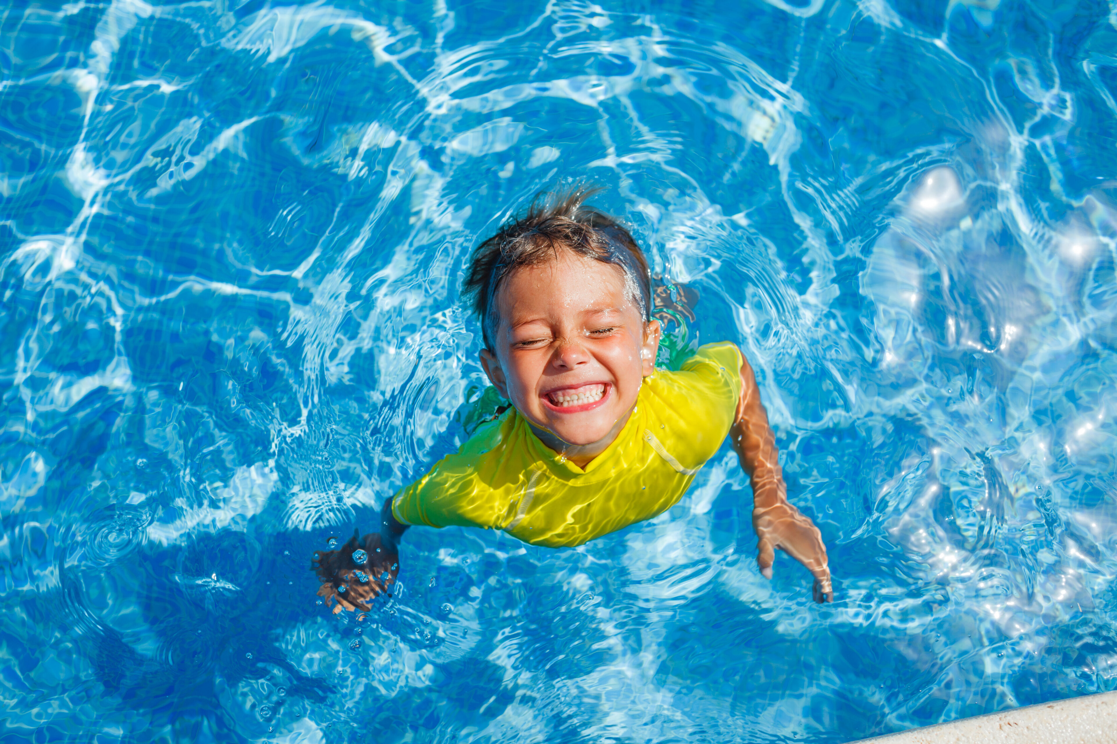 Child in a yellow shirt smiles with eyes closed while standing in a clear blue swimming pool, viewed from above.