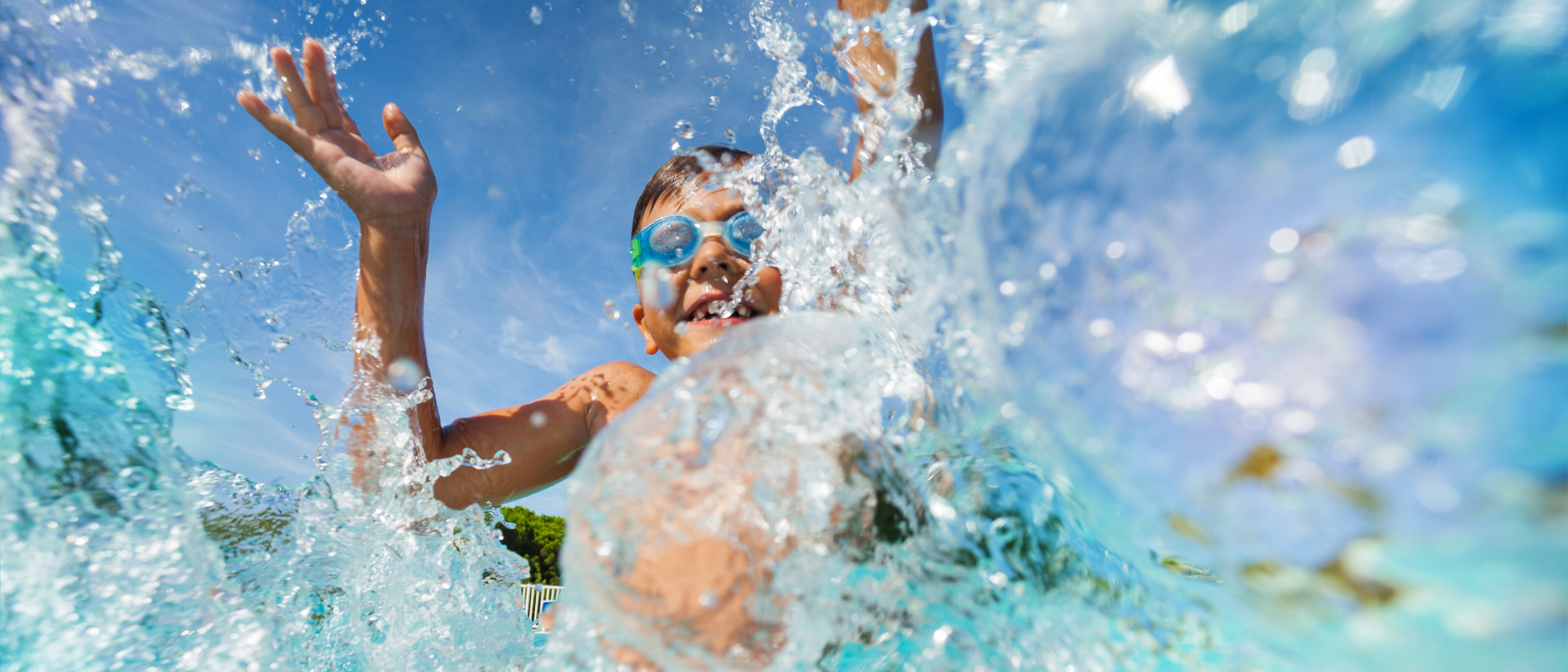 A child wearing goggles splashes in a pool, with water droplets in the foreground and a clear blue sky in the background.